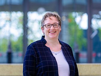Frauke Meyer, with short hair, jacket and glasses, stands in the "Glasshalle". In the background, you can see the green trees on campus.